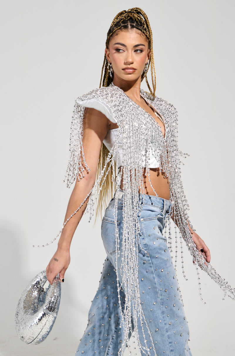 A woman with long braided hair embodies the essence of a party-perfect piece wearing the IN MY OWN LANE SILVER BEADED TOP paired with blue jeans accented with rhinestones. Holding a metallic silver sequined hat, she gazes confidently to the side against a plain white background.