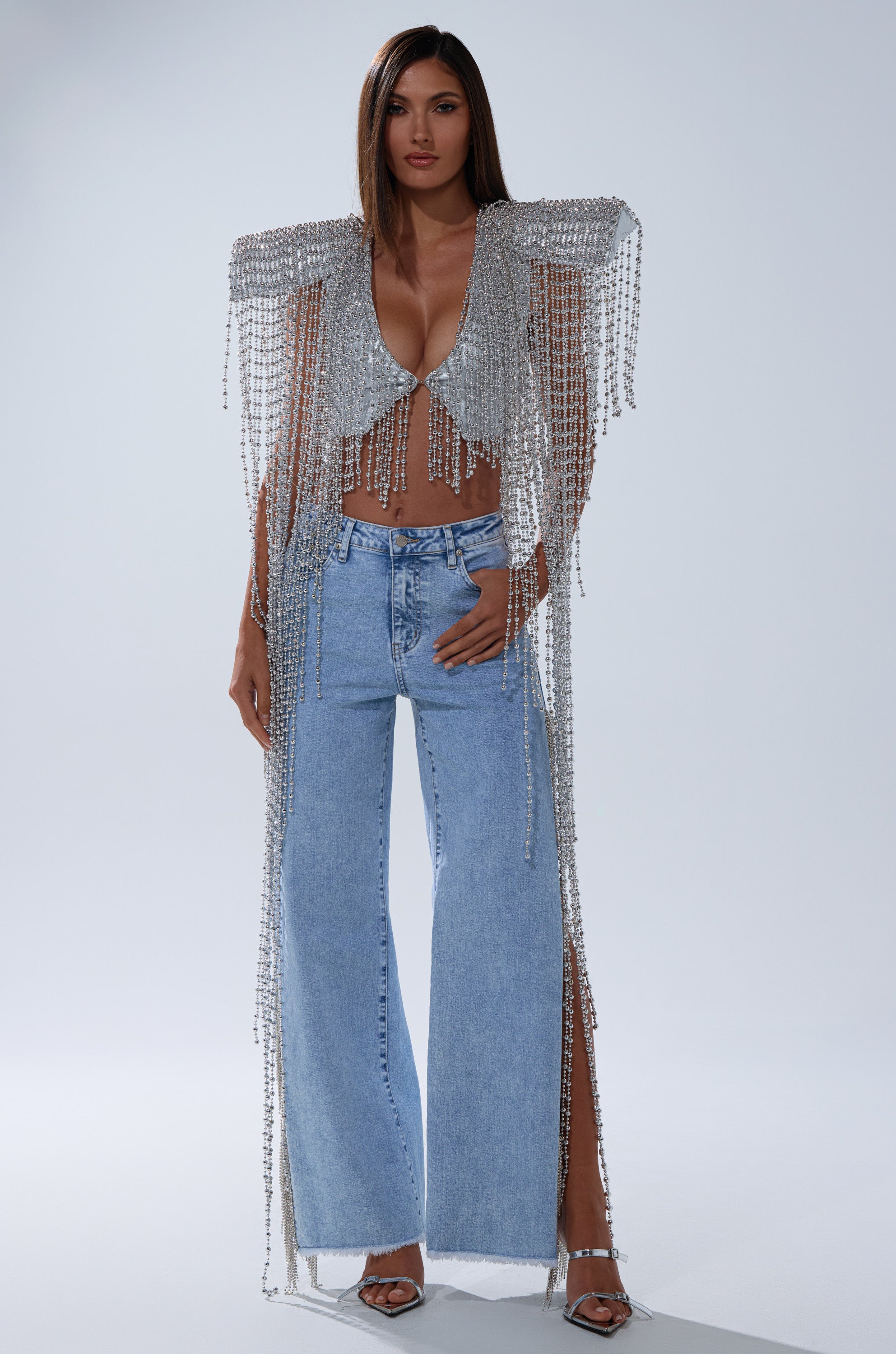 A woman poses confidently against a plain background wearing the IN MY OWN LANE SILVER BEADED TOP with shimmering beads and crystal fringe, styled with light blue wide-leg jeans and silver high-heeled sandals.