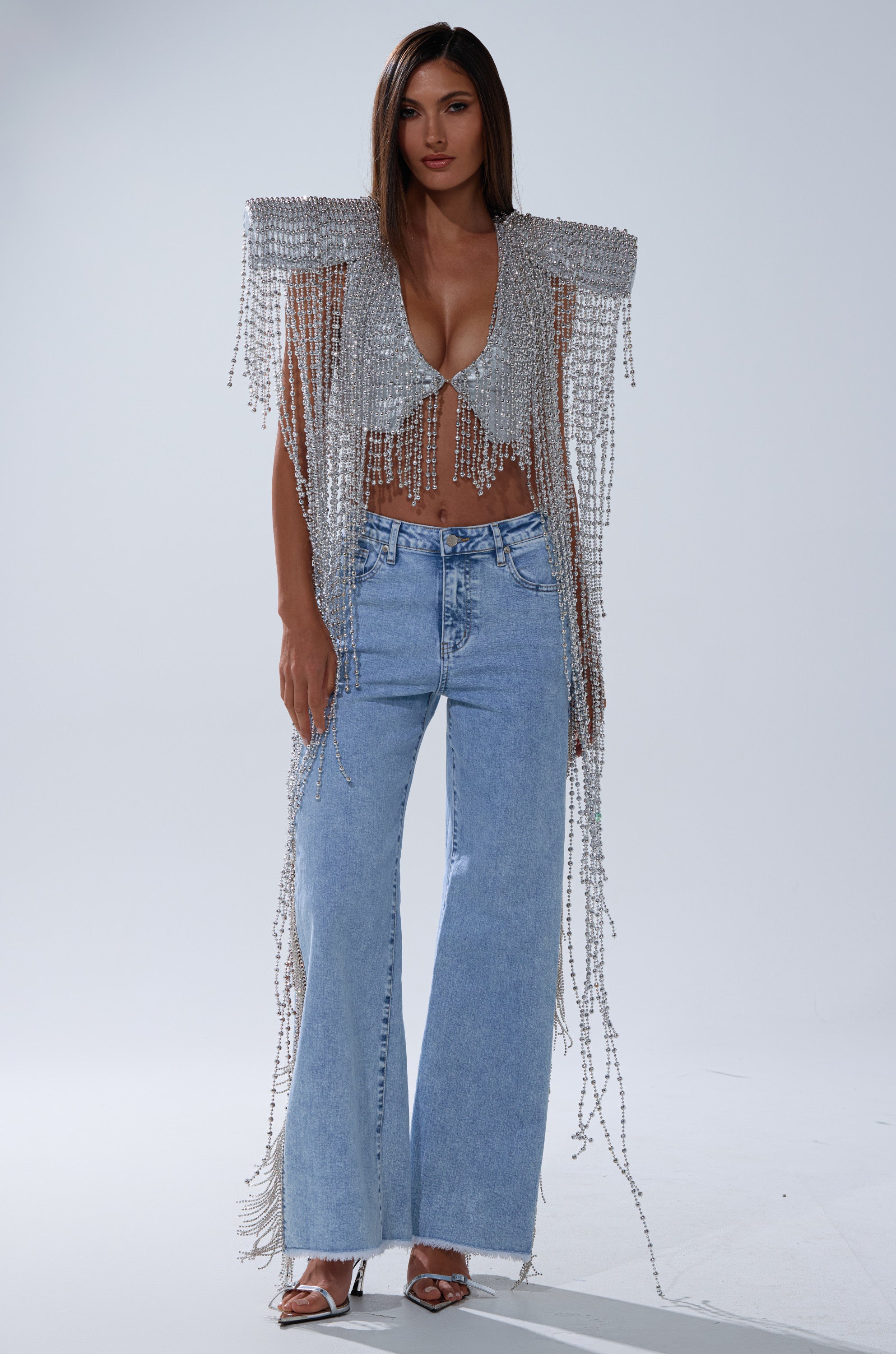 A woman stands wearing light blue wide-leg jeans, silver high-heeled sandals, and the IN MY OWN LANE SILVER BEADED TOP—a sparkling party top with dramatic fringe draping from the shoulders to the floor.