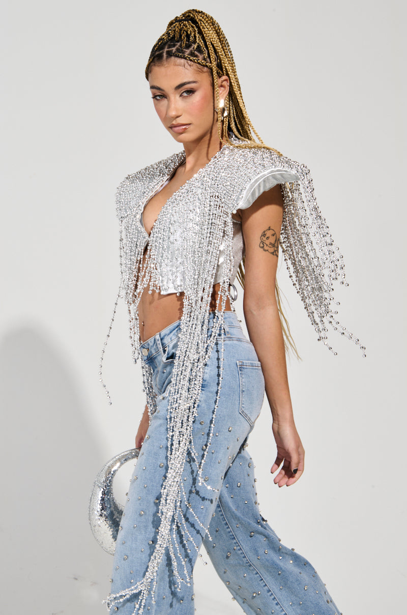 A person with braided hair dons the "IN MY OWN LANE SILVER BEADED TOP," a striking metallic silver sequin piece with long fringes, paired with pearl-embellished light blue jeans. They carry a small, chic bag and pose against a simple backdrop, showcasing the epitome of party-ready fashion.