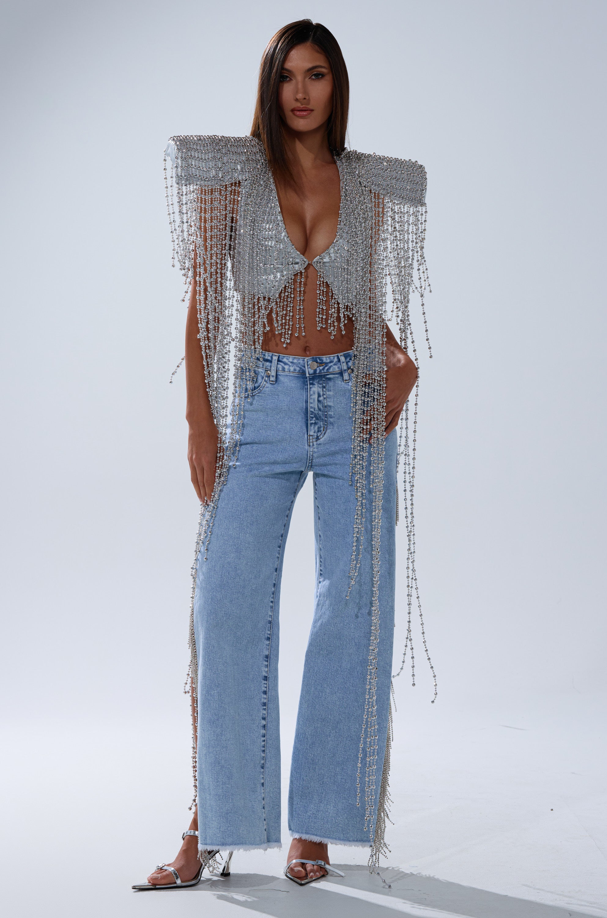 A woman poses in the IN MY OWN LANE SILVER BEADED TOP with beaded fringe on the shoulders and sleeves, paired with light blue wide-leg jeans and silver high heels, against a plain light background.