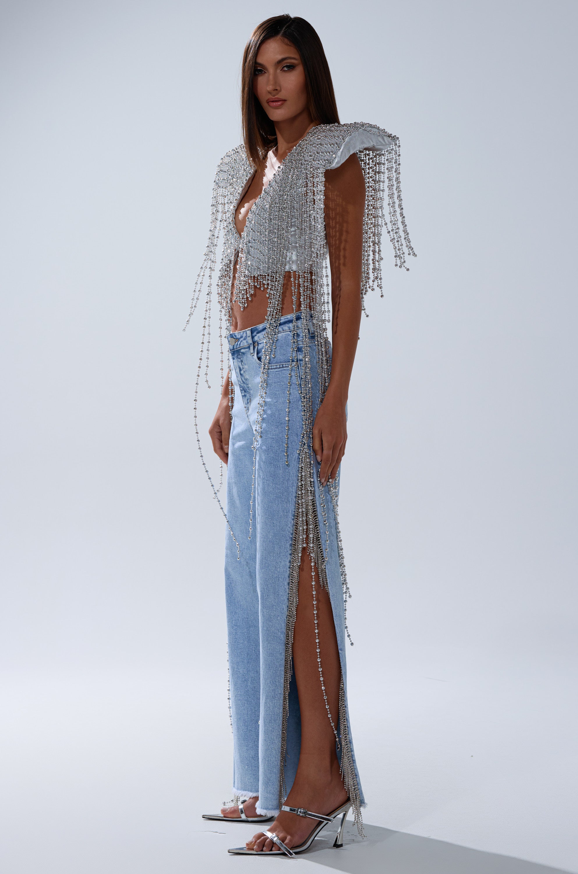 A woman with straight, dark hair models the IN MY OWN LANE SILVER BEADED TOP, featuring fringe and draped beads, paired with high-slit light blue jeans and metallic heels against a plain background.