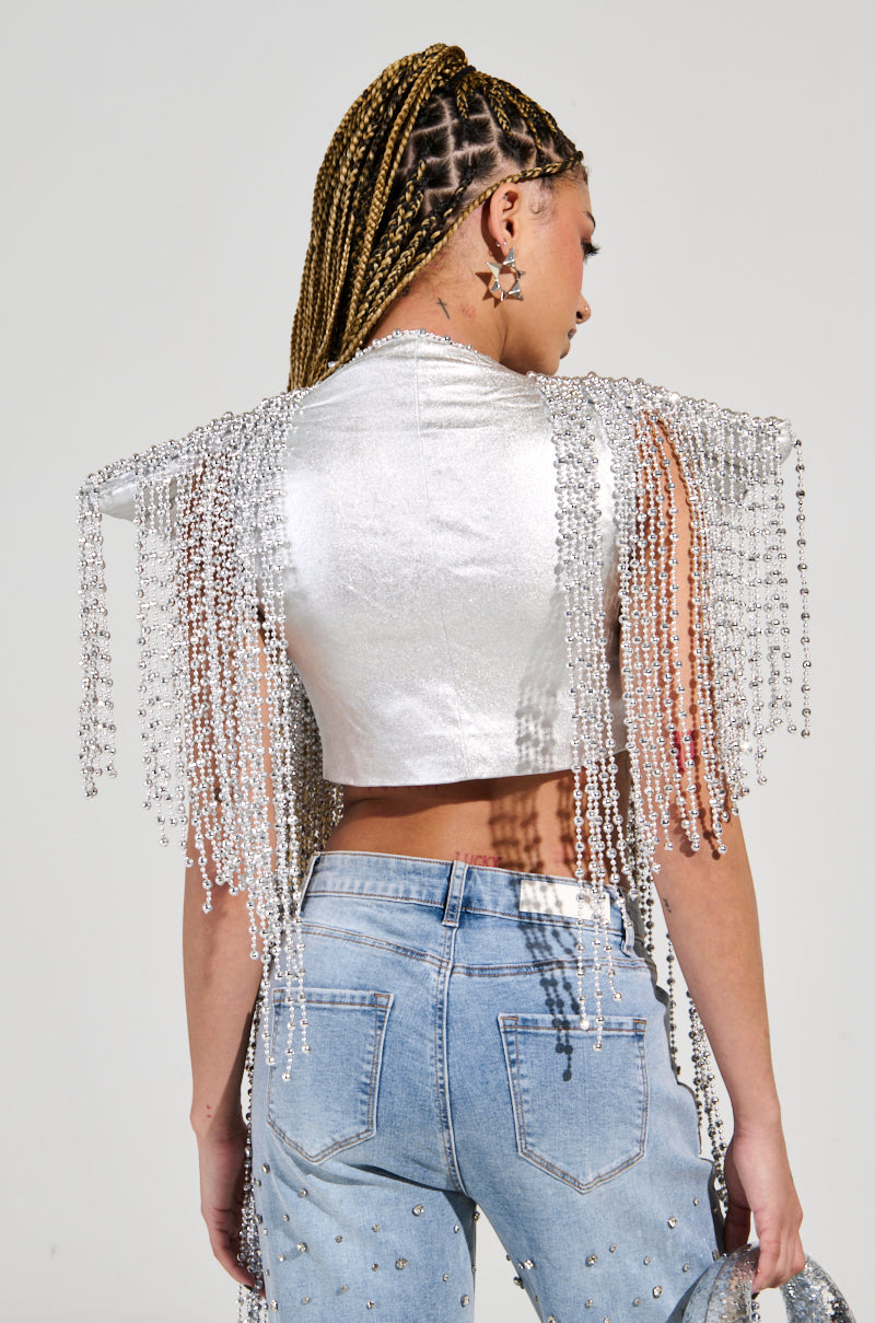A person with braided hair stands facing away, showcasing a party-perfect metallic silver top with beaded fringe on the shoulders—identified as the "In My Own Lane Silver Beaded Top"—and dangling star earrings. They pair this ensemble with light blue jeans adorned with decorative beadwork.