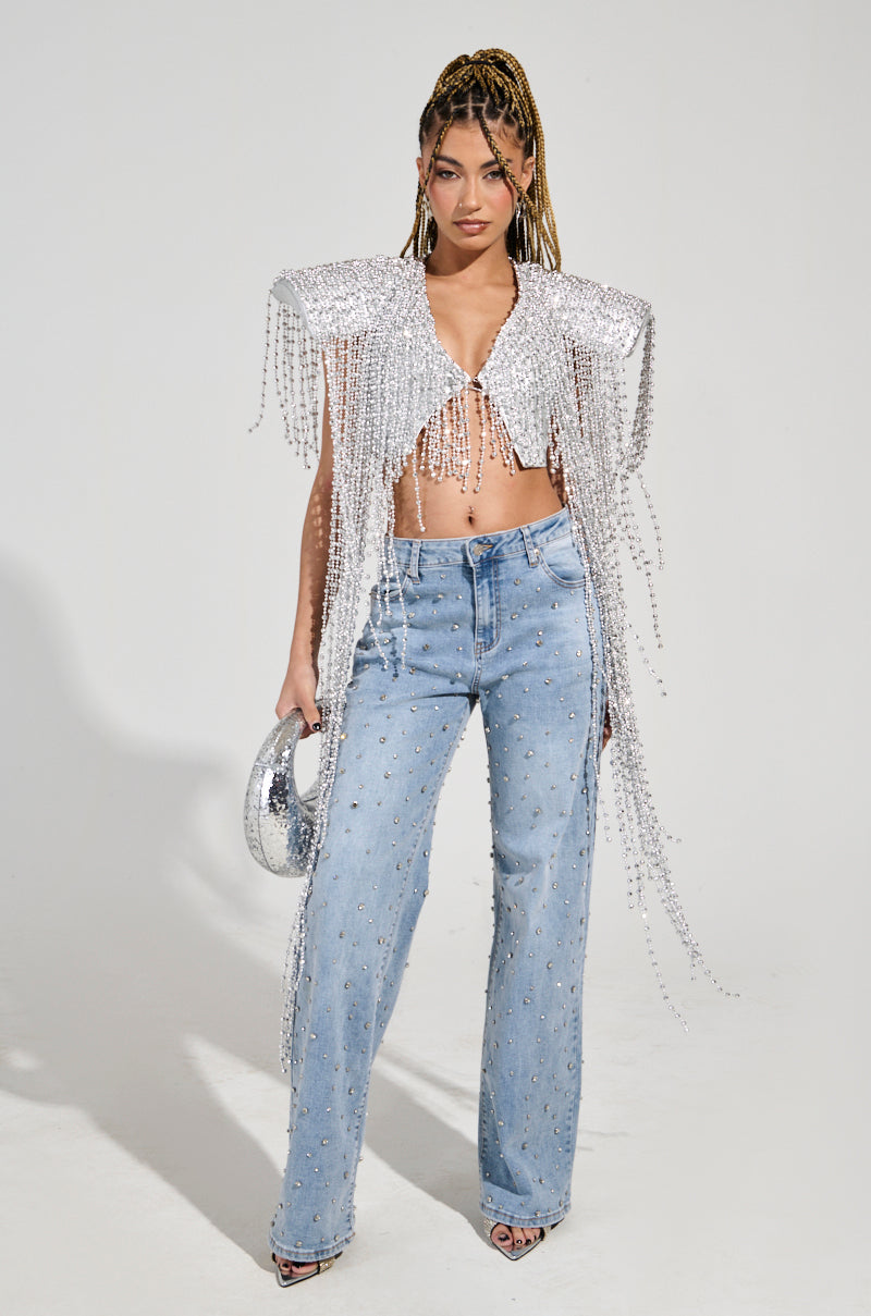 A person stands confidently wearing the party-perfect "IN MY OWN LANE SILVER BEADED TOP," styled over a bralette and high-waisted embellished blue jeans. They hold a silver bag and have long braided hair, with the plain white backdrop highlighting their outfit beautifully.