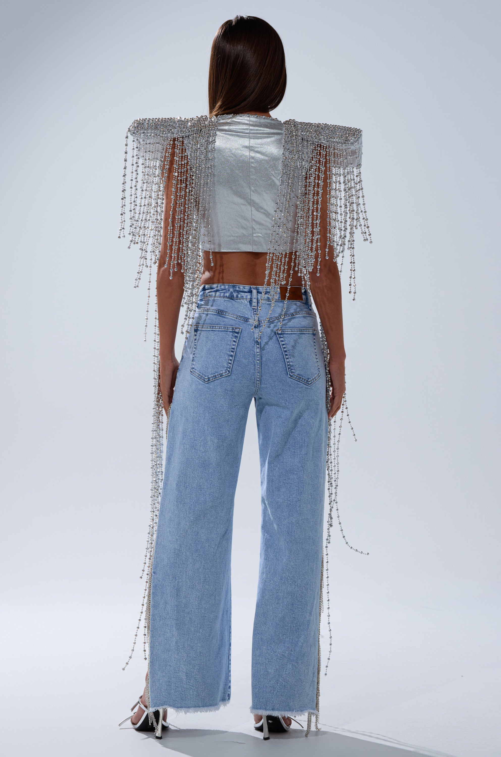 A person with long straight hair faces away, wearing the IN MY OWN LANE SILVER BEADED TOP with draped fringe shoulders, light blue high-waisted wide-leg jeans, and white heeled sandals.
