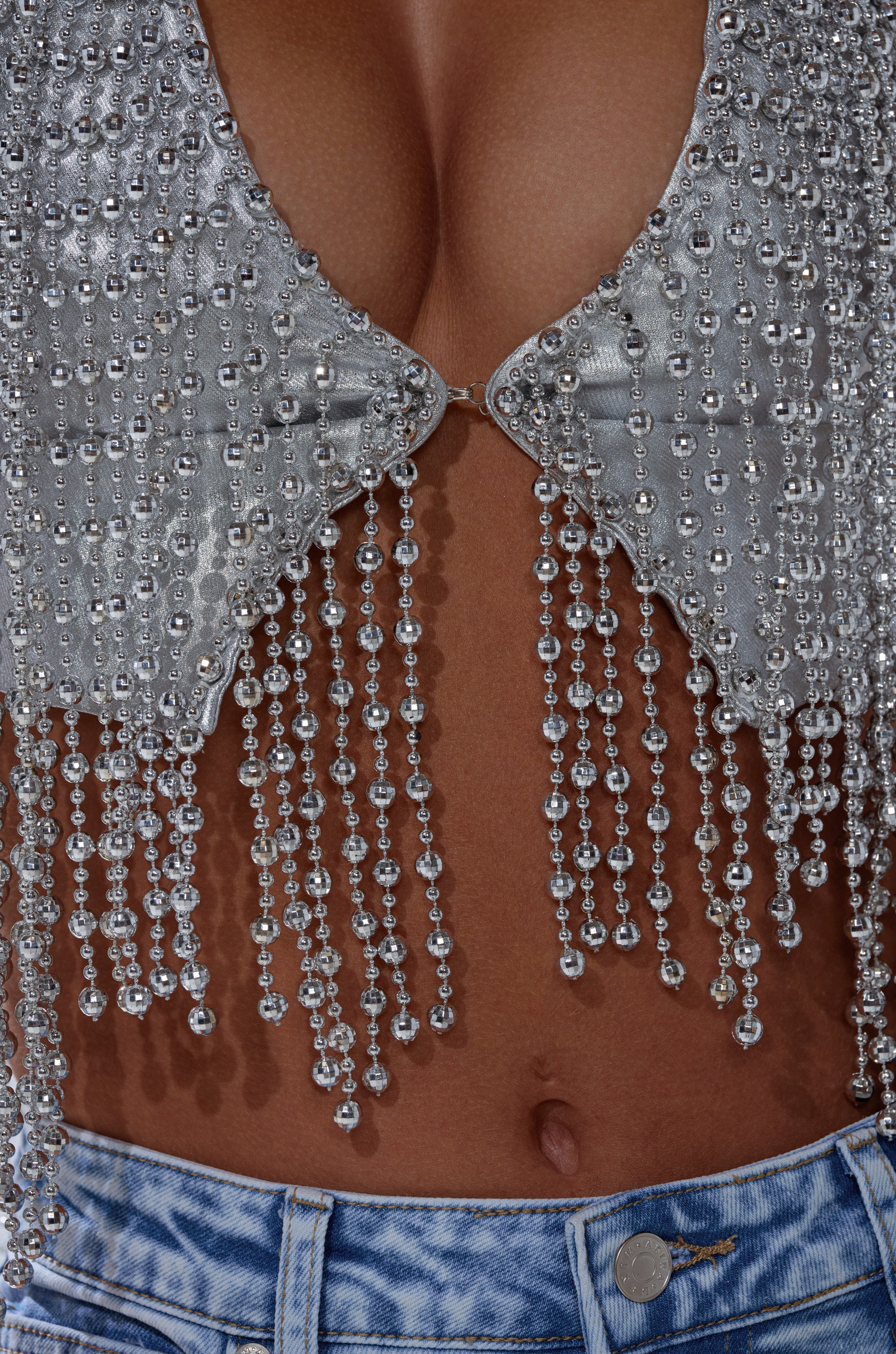 Close-up of a person wearing the IN MY OWN LANE SILVER BEADED TOP—a sparkling, silver beaded crop with dangling crystal strands—paired with high-waisted, light blue acid-wash jeans. The chic top reveals part of the midriff and cleavage.