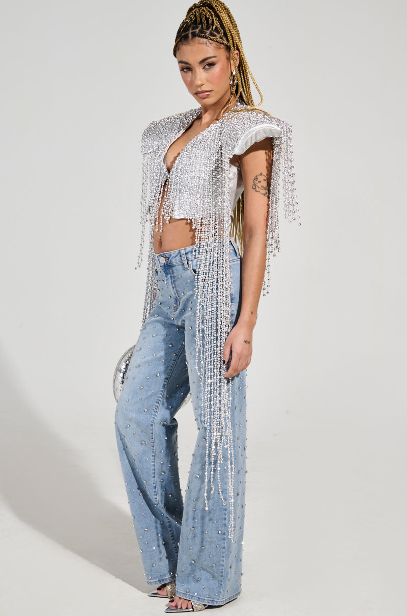 A person with long, braided hair stands confidently in the IN MY OWN LANE SILVER BEADED TOP, adorned with fringe and perfectly catching the light as the ultimate party-perfect piece. Paired with light blue jeans and rhinestones, their stylish attire is highlighted against the plain background.
