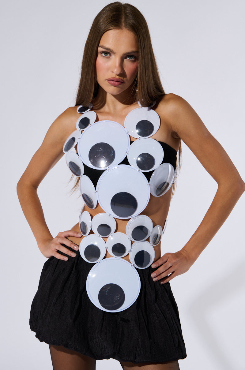 A woman with long straight hair wears a quirky GOOGLY EYED SLEEVELESS FASHION TOP, which is adorned with numerous large plastic googly eyes in a unique pattern covering her chest and midriff. She pairs this eye-catching top with a black ruffled skirt. Posing with her hands on her hips against a plain background, she showcases the distinctive style of the top.