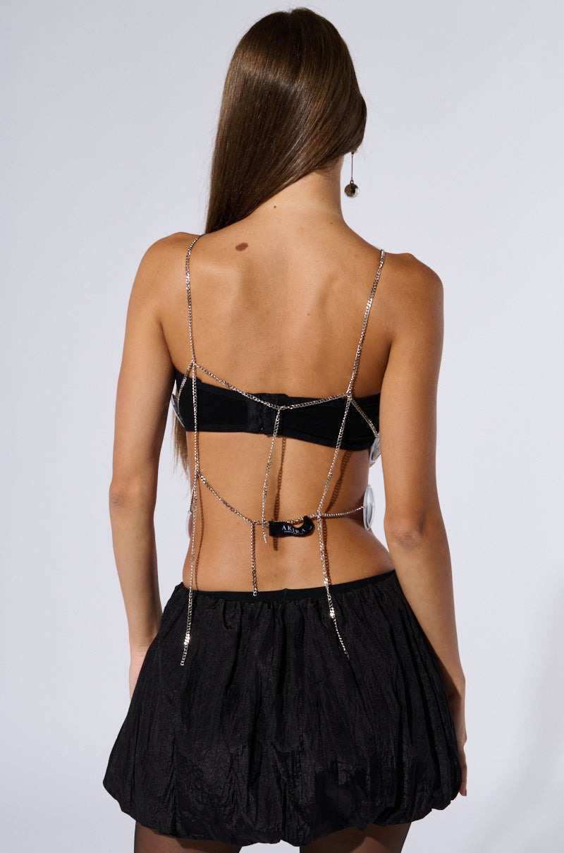A woman with long brown hair is facing away, showcasing the GOOGLY EYED SLEEVELESS FASHION TOP, a black bralette featuring metallic silver chain link straps and intricate back detailing. She pairs it with a black, textured skirt that reaches mid-thigh. The background is plain and light-colored.