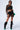 A person wearing a stylish outfit consisting of a sheer top and the LATE NIGHT VIBES MINI SKIRT adorned with black geometric diamonds, paired with knee-high scrunched black boots with green tips. They carry a green handbag and wear sunglasses, posing confidently.