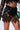 Close-up shot of a person's midsection wearing a black, geometric cut-out top and the LATE NIGHT VIBES MINI SKIRT, crafted from stretchy faux leather. The ensemble boasts an edgy, reflective diamond pattern. Visible tattoos adorn their torso and thigh as they hold a green handbag.