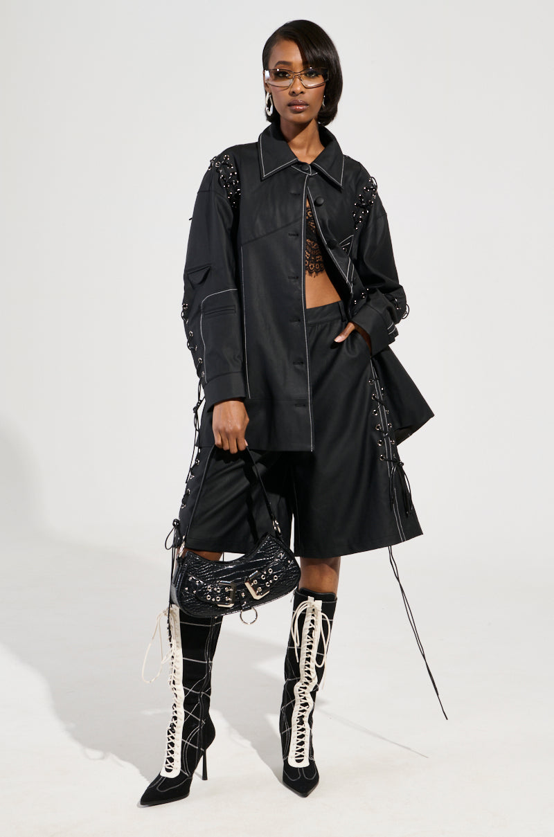A person confidently poses in a chic black ensemble, highlighted by the COLD HEART SHORT—faux leather shorts with contrast stitching—and an oversize jacket. They accessorize with a small handbag while wearing sunglasses and knee-high boots adorned with intricate white laces, standing against a light background.
