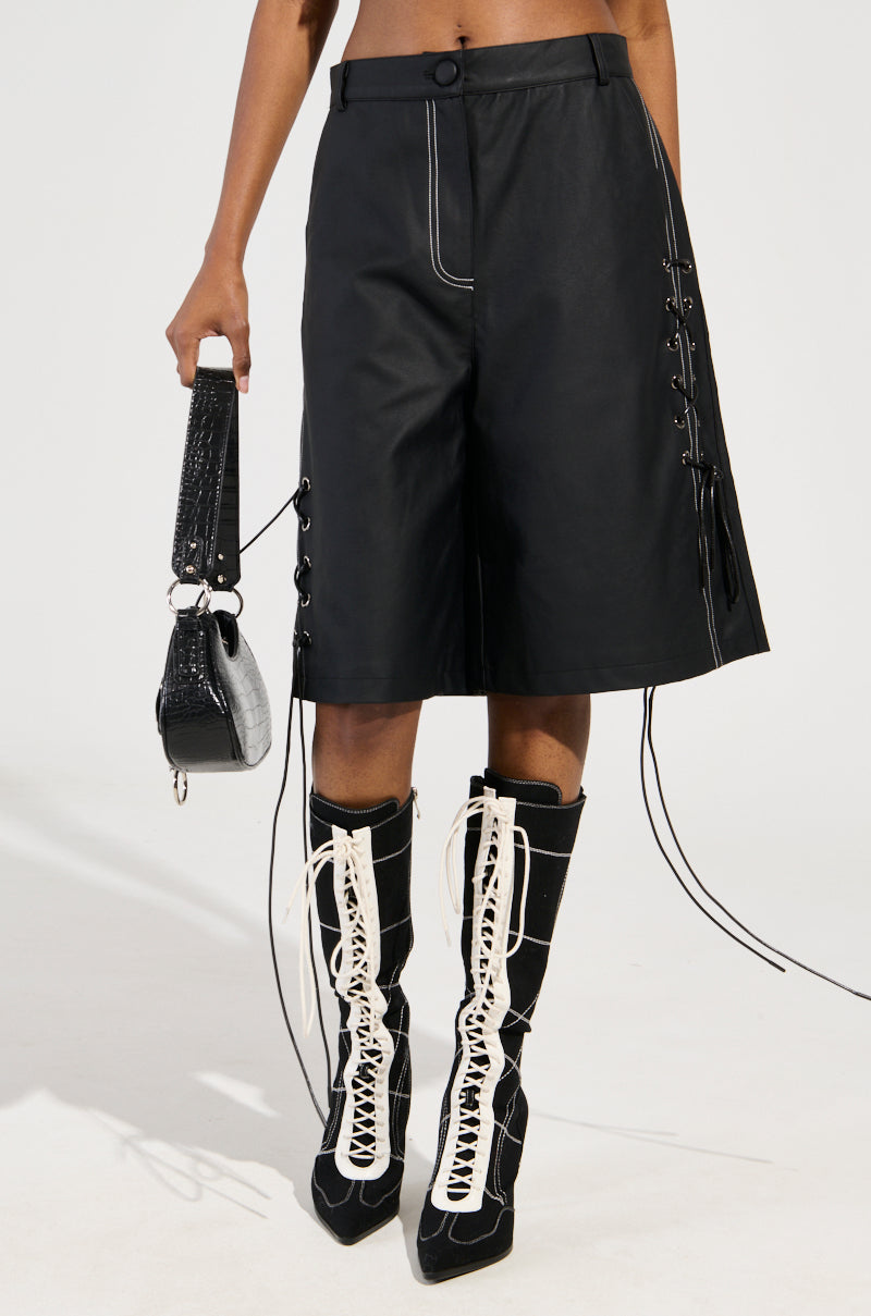 A person is dressed in the COLD HEART SHORT, a black skirt featuring contrast stitching and lace-up sides, complemented by tall black and white lace-up boots. They carry a stylish black and gray handbag against a plain, light-colored background.