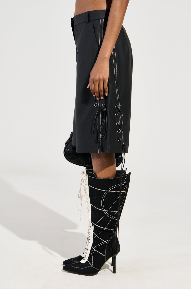 A person is seen in profile wearing knee-high black boots with white laces and black knee-length faux leather shorts called COLD HEART SHORT, featuring decorative contrast stitching on the side, set against a plain white background.