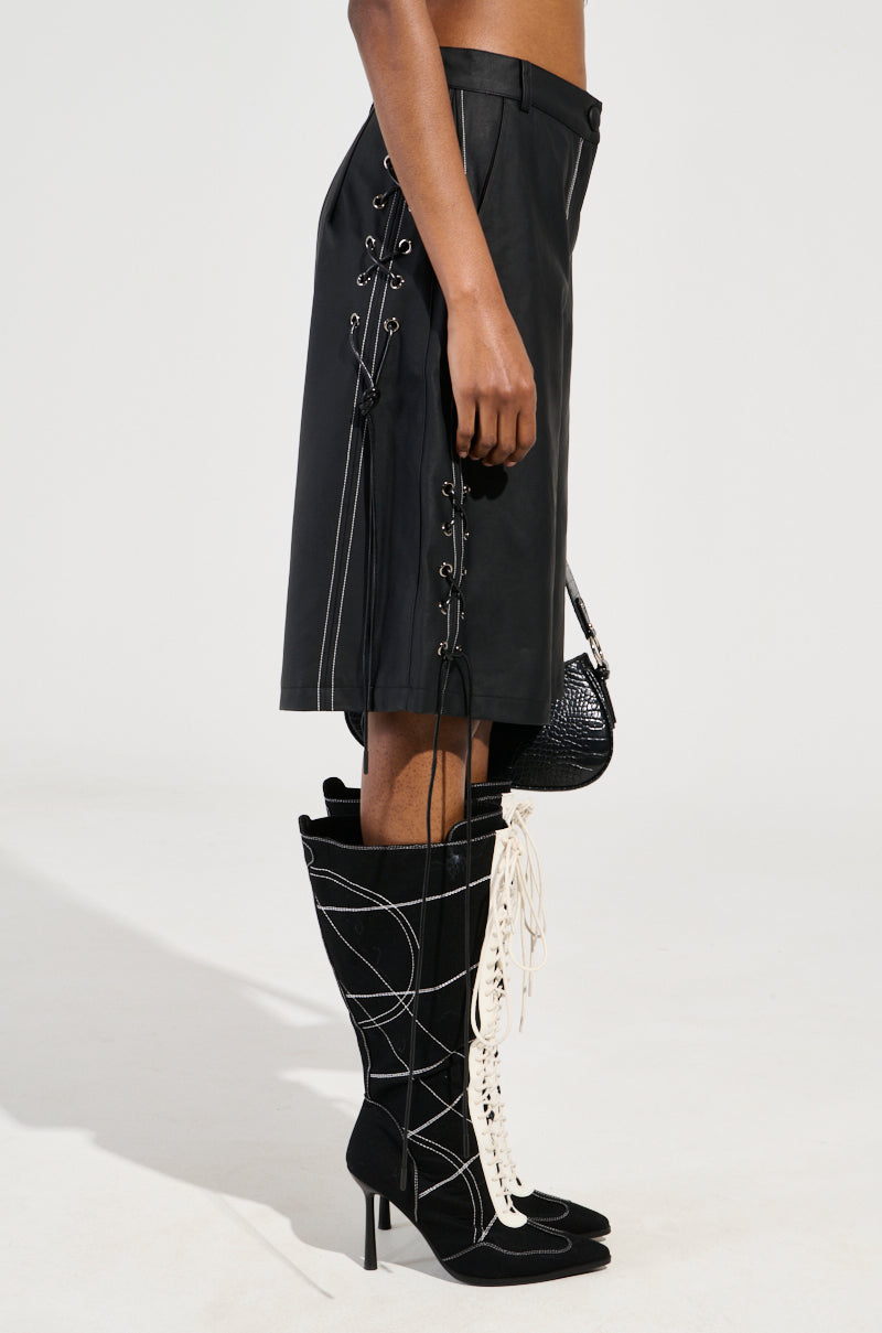 A person wearing the COLD HEART SHORT, a knee-length black leather skirt with lace-up details on the side, is shown in profile. They are also sporting black high-heeled boots featuring white contrast stitching and carrying a small black textured handbag.