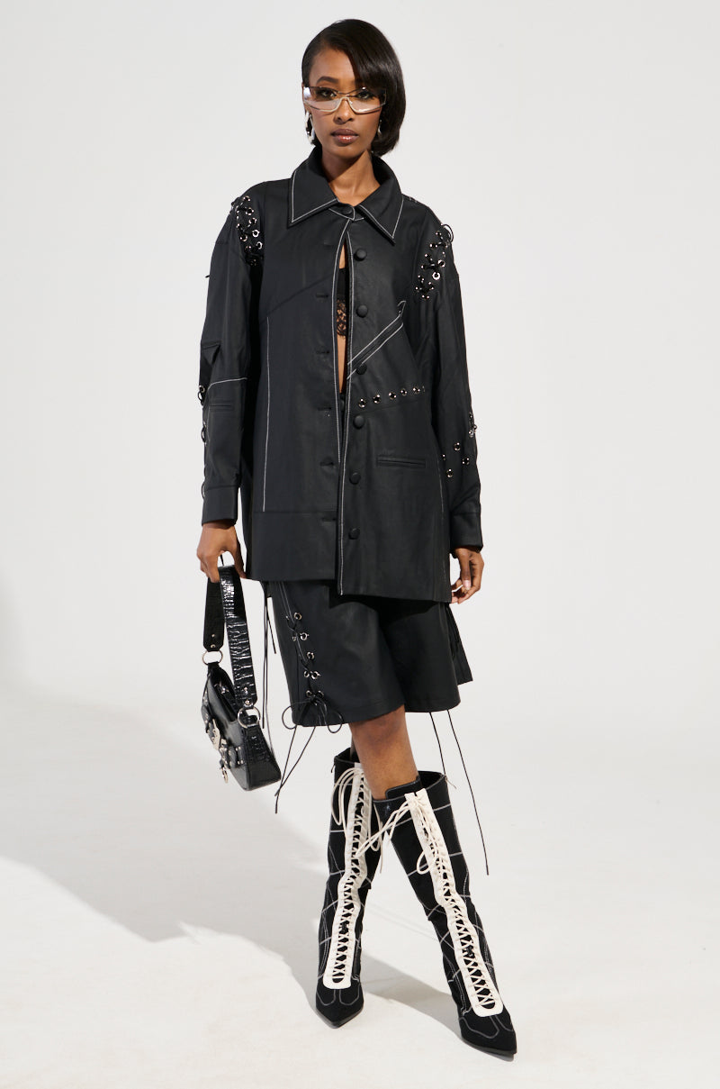 A person stands confidently in a stylish black outfit with unique details, including a jacket and skirt adorned with eyelets and laces. Their COLD HEART SHORTS in faux leather display contrast stitching, adding flair. Knee-high boots feature intricate lace-up designs. They hold a matching handbag and wear sunglasses.