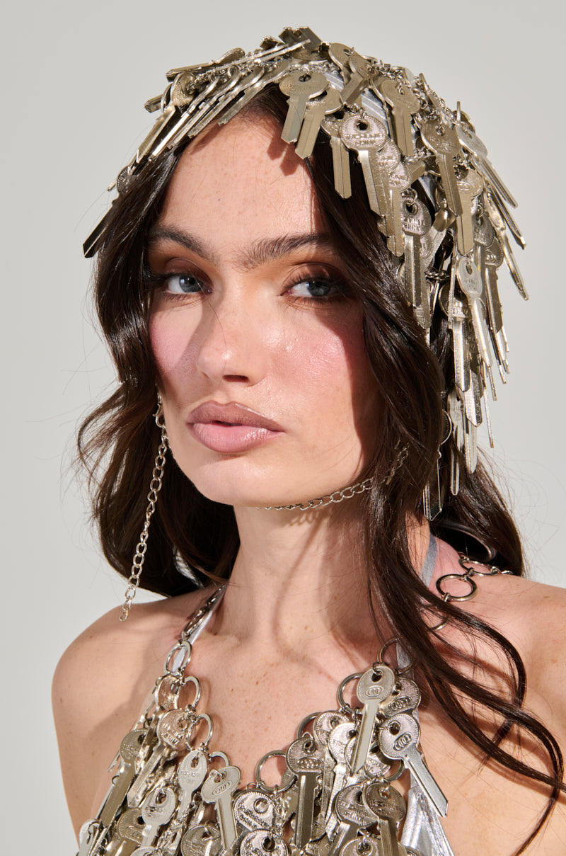 A woman with long brown hair wears the distinctive YOU HAVE THE KEY HAIR PIECE, crafted from silver keys as part of the exclusive AKIRA Label. She complements it with a matching necklace and gazes slightly to the side against a neutral, light background.