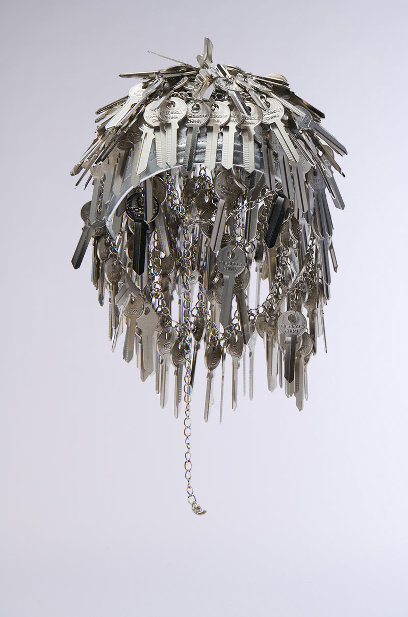 A chandelier made from numerous silver keys, reminiscent of the YOU HAVE THE KEY HAIR PIECE by AKIRA Label, hangs in a dome shape against a plain background. A chain elegantly dangles from the center, enhancing the intricate and artistic design. 5