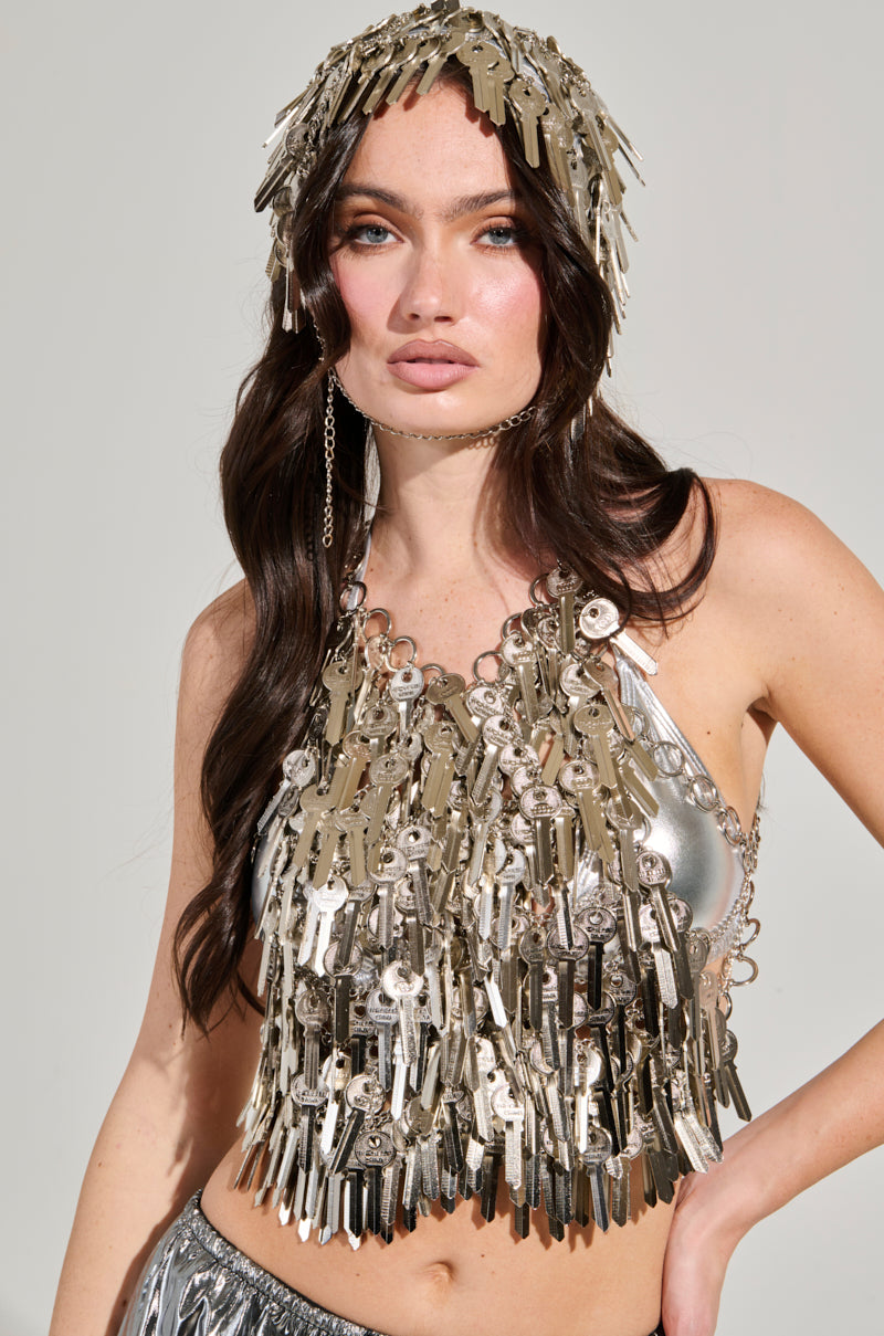 A woman showcases a unique outfit made of metallic keys, featuring a silver keys top and a YOU HAVE THE KEY HAIR PIECE by AKIRA Label adorned with numerous keys. She's dressed in shimmering metallic fabric against a plain background, emphasizing her distinctive attire.