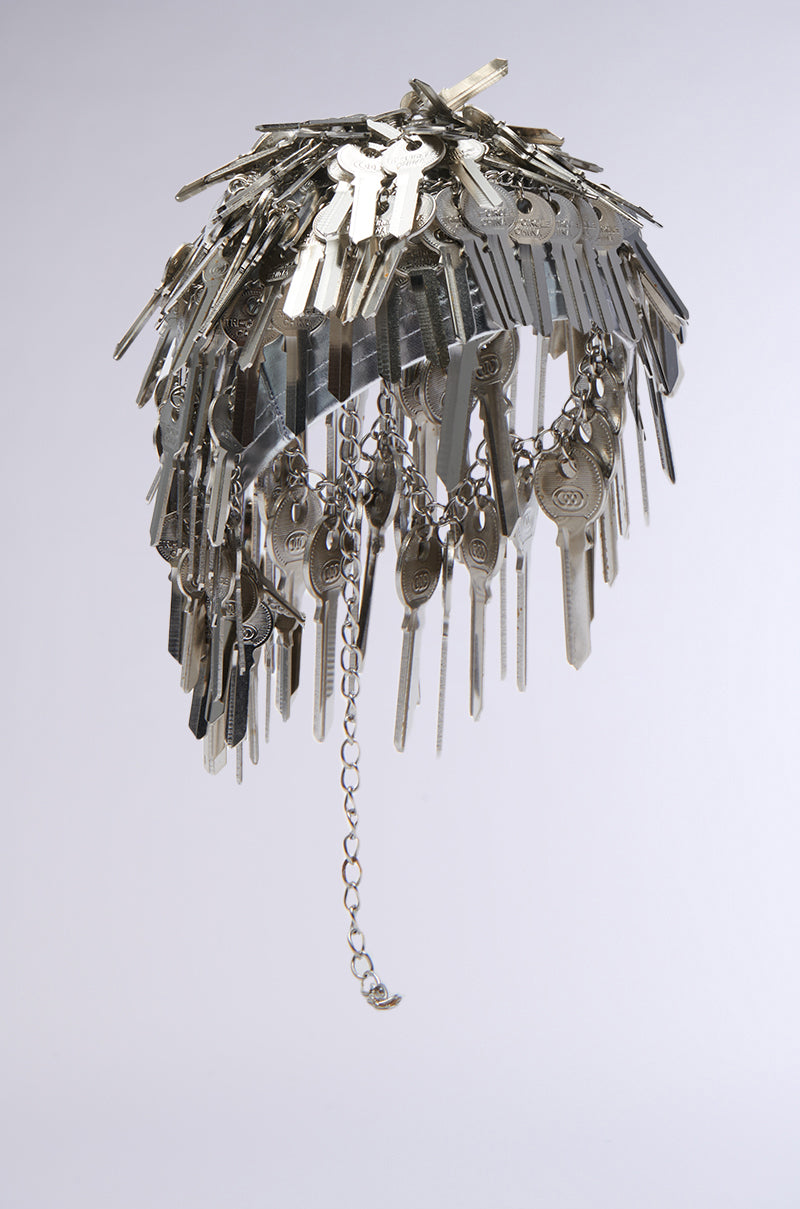 A hanging sculpture resembling an umbrella, crafted entirely from silver keys, dangles gracefully, reminiscent of the intricate designs seen in the YOU HAVE THE KEY HAIR PIECE collections, with a single chain beautifully cascading from its center.
