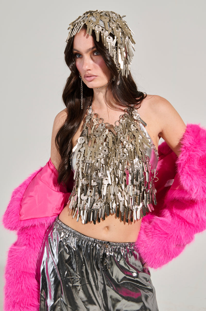 A woman wears a top and the YOU HAVE THE KEY HAIR PIECE, posing confidently in an AKIRA Label creation. Her long, dark hair cascades over a pink fur coat draped across her shoulders, perfectly complementing her metallic silver pants against the plain gray backdrop.