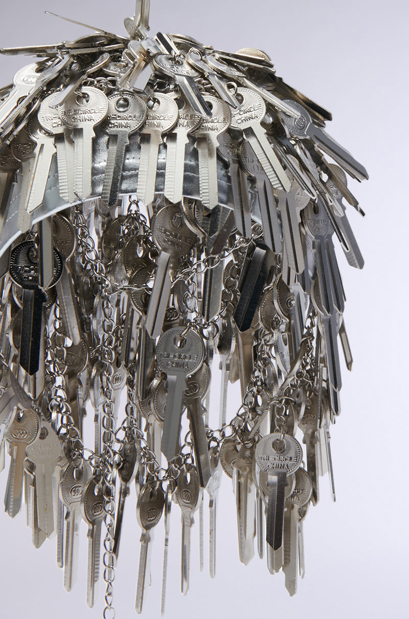 A hair piece crafted from numerous silver keys hanging on chains, designed in a circular, tiered fashion reminiscent of an elegant chandelier. The detailed arrangement stands out against the plain white background, evoking the essence of an exquisite YOU HAVE THE KEY HAIR PIECE from the AKIRA Label. 7