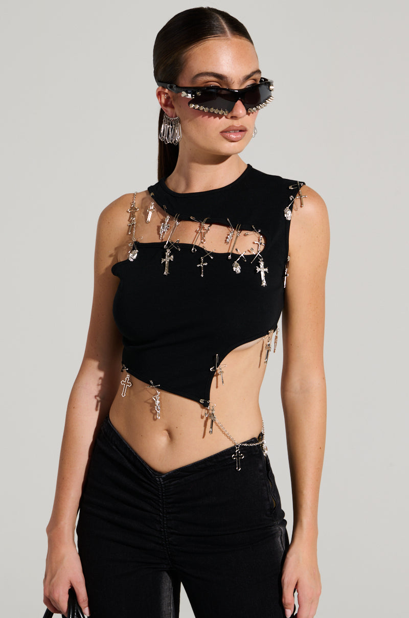 A person with long hair sports cat-eye sunglasses and a sleeveless black AKIRA Label BLESSED TOP adorned with metal crosses and safety pins. They complete their edgy outfit with black pants and hoop earrings, standing out against a plain background with a rebellious flair.
