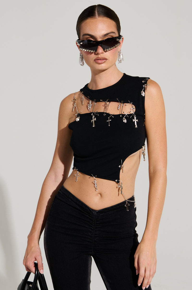 A woman dressed in a BLESSED TOP paired with futuristic sunglasses and large hoop earrings. The top, featuring cross-shaped chain details, complements her confident pose as she holds a black bag against a neutral backdrop.