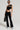 A person wearing an AKIRA Label BLESSED TOP featuring an asymmetrical neckline and cut-outs, paired with black flared pants and light blue shoes, poses confidently against a plain background. They hold a black handbag and sport stylish sunglasses and earrings.