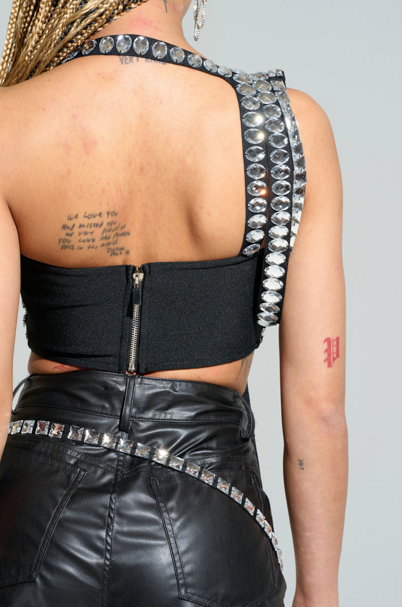 A person with braided hair is wearing the KIMMY ASYMMETRICAL RHINESTONE TOP in black, featuring silver studs, paired with a matching black leather skirt. The ensemble is enhanced by metallic back zippers that lend an edgy flair, and tattoos are visible on the person's back and arm.