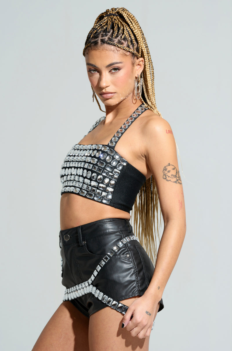 A person with long braided hair poses against a plain background, dressed in the KIMMY Asymmetrical Rhinestone Top paired with black leather shorts. They flaunt tattoos on one arm, accessorized with earrings, achieving an edgy appearance highlighted by gemstone detailing.
