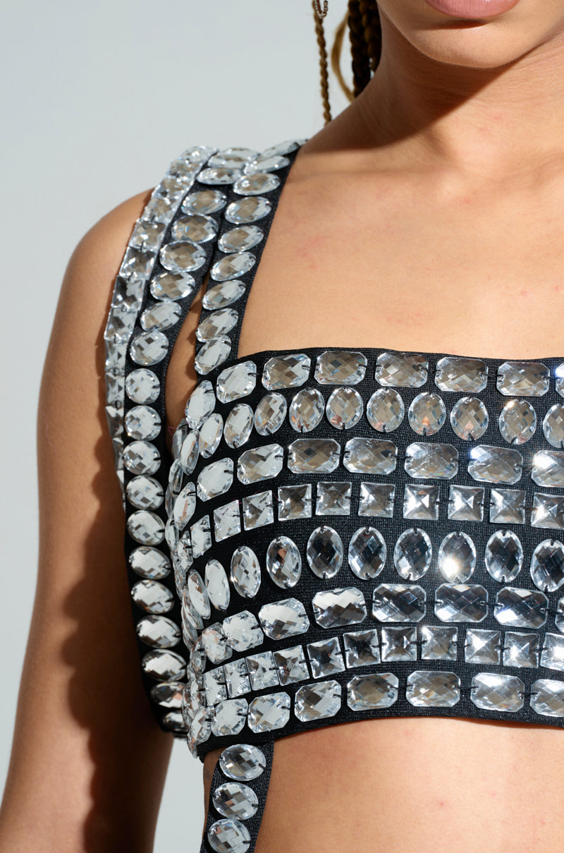 Close-up of someone wearing the KIMMY ASYMMETRICAL RHINESTONE TOP, embellished with multiple rows of large, shiny gemstones. The top features broad straps and a stretchy base, allowing the oval and square-shaped rhinestones to beautifully reflect light across the upper torso.