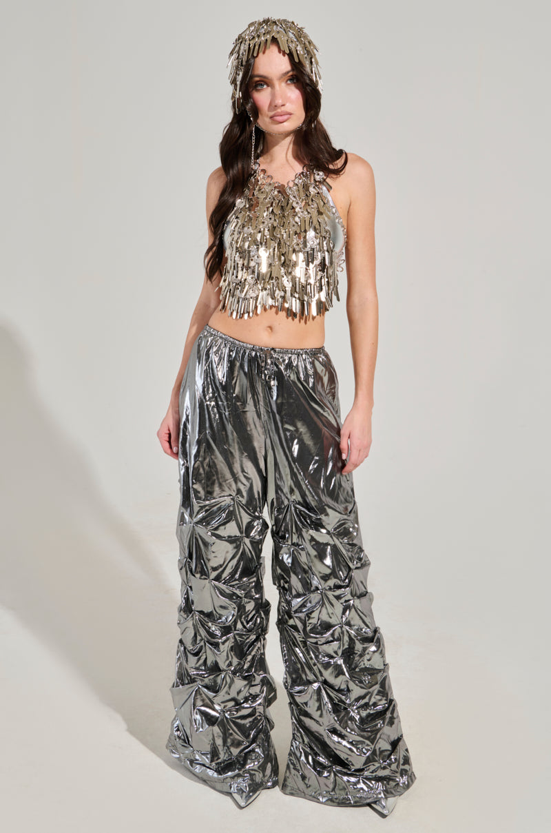 A person wearing the UNDER LOCK AND KEY TOP, a dazzling metallic silver piece, paired with crinkled pants and a shimmering silver headpiece, stands against a plain background. The outfit is reflective and eye-catching.