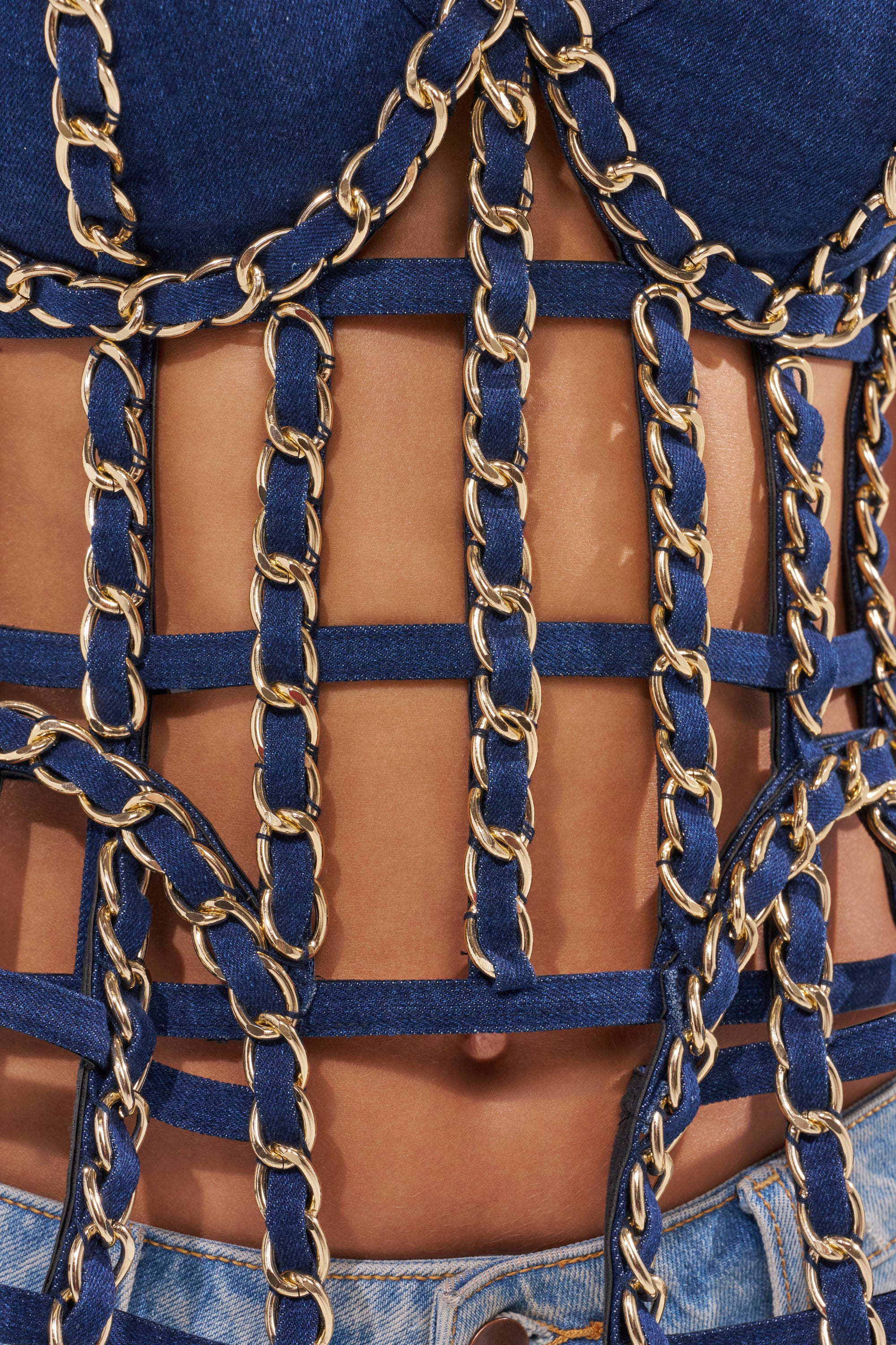 Close-up of a person wearing the DOWN SOUTH CHAIN DETAIL BUSTIER TOP, a blue denim piece with gold chain grid detailing that reveals skin below. The waistband of blue denim jeans is visible, showcasing avant-garde fashion. 8