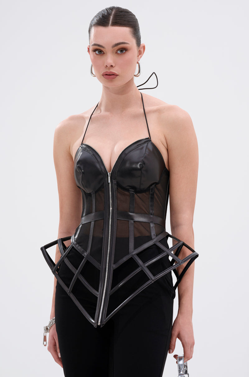 A woman wearing the ALL THE DRAMA CUT OUT CORSET—a black structured top with sheer mesh panels, thin halter straps, and black pants—stands against a light background. Her hair is slicked back with a neutral expression.