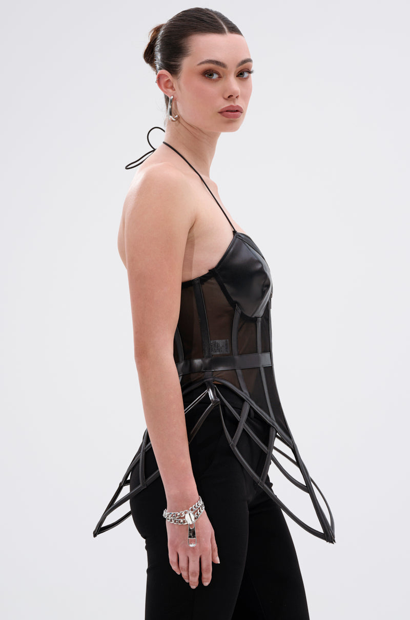 A woman with dark hair in a sleek updo models the ALL THE DRAMA CUT OUT CORSET, featuring cage-like structured details, paired with black pants. She poses in profile against a plain light gray background.