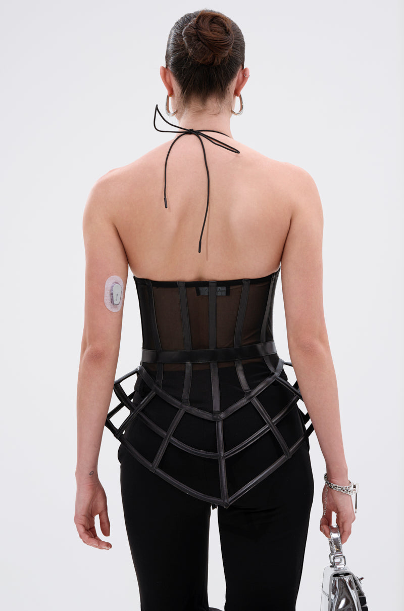 A woman with dark hair in a bun is photographed from behind wearing the ALL THE DRAMA CUT OUT CORSET, featuring structured cage detailing and sheer mesh over black pants, holding a silver purse. An adhesive medical device is visible on her upper left arm.