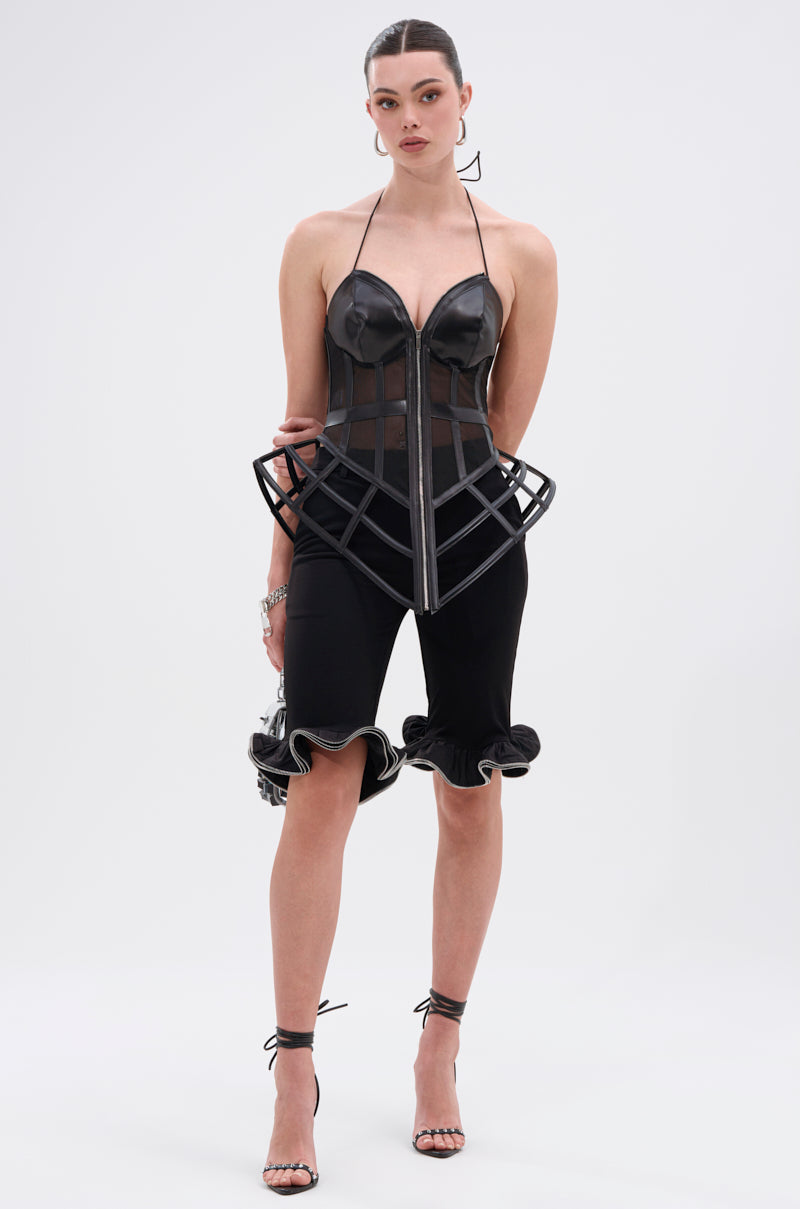 A woman exudes confidence in the ALL THE DRAMA CUT OUT CORSET, featuring black faux leather, sheer mesh panels, and a structured cage design at the hips. She pairs it with black ruffled knee-length shorts and strappy heels against a plain light backdrop.