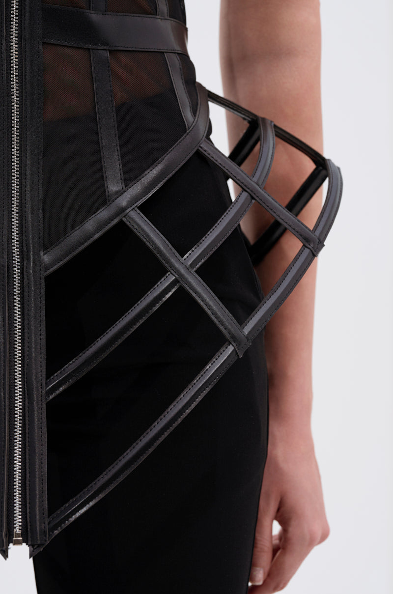 A close-up of a person wearing black pants and the ALL THE DRAMA CUT OUT CORSET, which features cage-like, boned panels extending outward at the hips. The background is plain and light-colored.