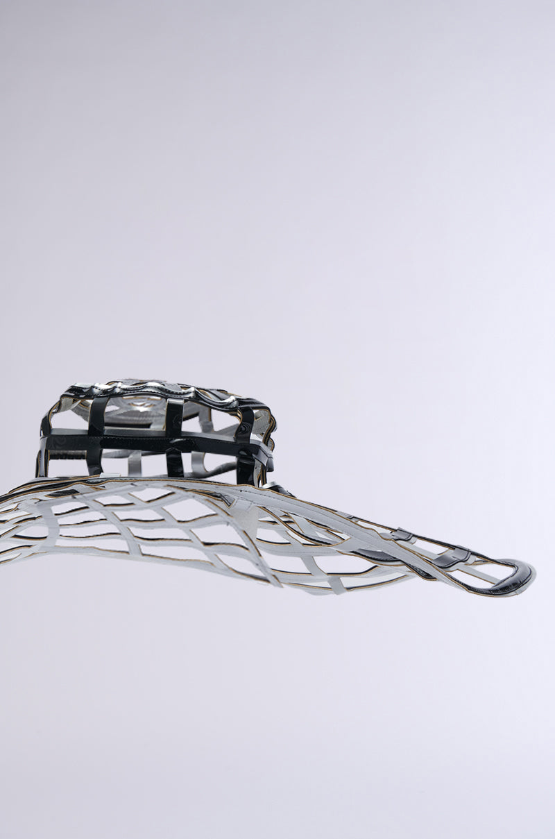 The IM OFF THE GRID BLACK SILVER HAT, a futuristic and sculptural accessory with intricate lattice-like patterns and a metallic finish, is displayed with its oversized brim suspended against a neutral gray background.