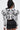 A woman with long dark hair, seen from behind, wears the white FRIEDA PUFF LONG SLEEVE CROPPED BLOUSE with bold black swirly patterns and 3D piping, paired with high-waisted black shorts.
