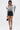 A woman poses against a white background wearing the FRIEDA PUFF LONG SLEEVE CROPPED BLOUSE with bold black swirl patterns, black high-waisted shorts, strappy heels, and a small black handbag. The blouse has a metallic back zipper closure.
