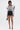 A woman stands against a white background wearing the FRIEDA PUFF LONG SLEEVE CROPPED BLOUSE, black high-waisted shorts, black strappy heels, and holding a small black handbag.