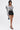 A woman stands against a plain white background wearing the FRIEDA PUFF LONG SLEEVE CROPPED BLOUSE with bold black piping, high-waisted black shorts, black heeled sandals, and a small black handbag.