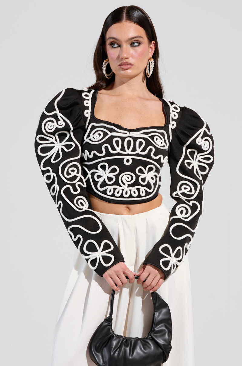 A woman with long dark hair wears a BLURRED LINES LONG SLEEVE BLOUSE, showcasing its dramatic sleeves and abstract design. She holds a small black handbag and stands against a light gray background, gazing sideways with a neutral expression.