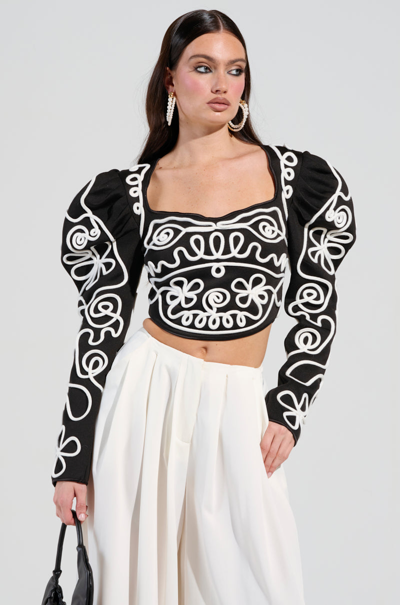 A woman with long dark hair is wearing the BLURRED LINES LONG SLEEVE BLOUSE, showcasing puffed sleeves and bold abstract patterns. She stylishly pairs it over a white skirt and complements her look with a black handbag and large earrings, creating a chic ensemble.