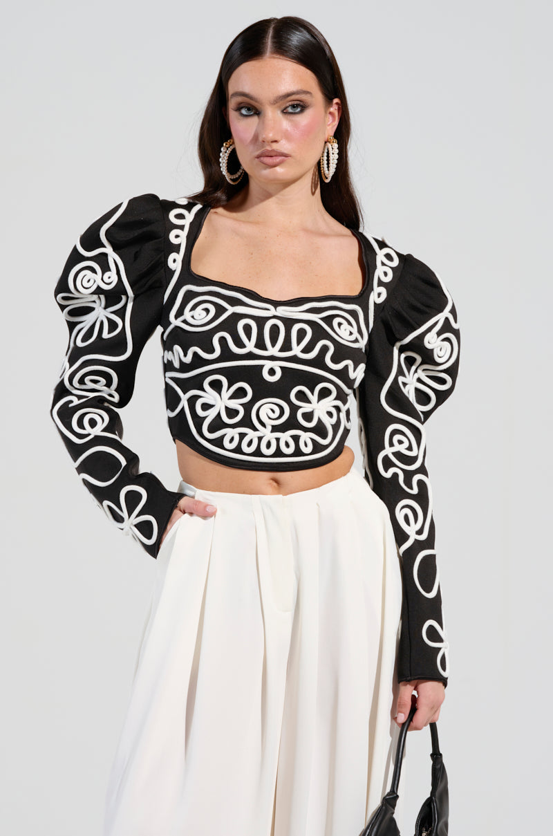 A woman with long dark hair models the Blurred Lines Long Sleeve Blouse in a striking black and white abstract pattern, featuring puffed sleeves. She pairs it elegantly with white wide-legged pants and stands against a light gray background. Her look is accessorized with a chic black handbag and large hoop earrings.