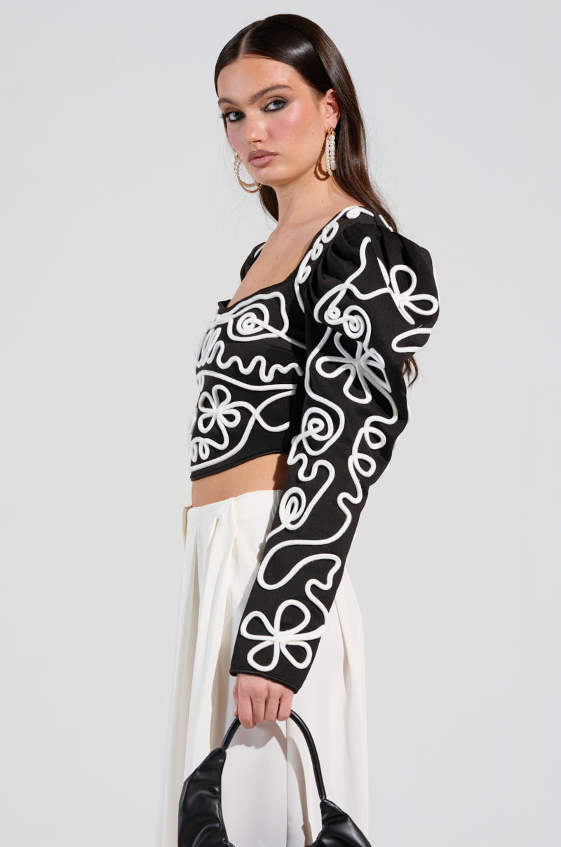 A woman wearing a BLURRED LINES LONG SLEEVE BLOUSE with white abstract patterns and white pants holds a black handbag. She has long dark hair and hoop earrings, posing against a plain light background.