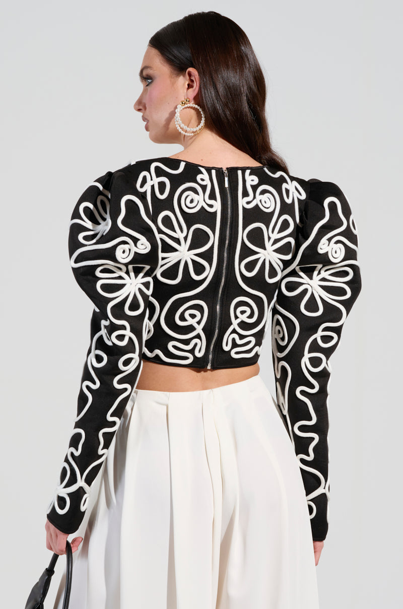 In the side and back view, an individual with long dark hair presents a striking look featuring the BLURRED LINES LONG SLEEVE BLOUSE, characterized by its abstract pattern and puffy shoulders, complemented by large circular earrings. This chic appearance is elegantly paired with high-waisted, wide-leg white pants.