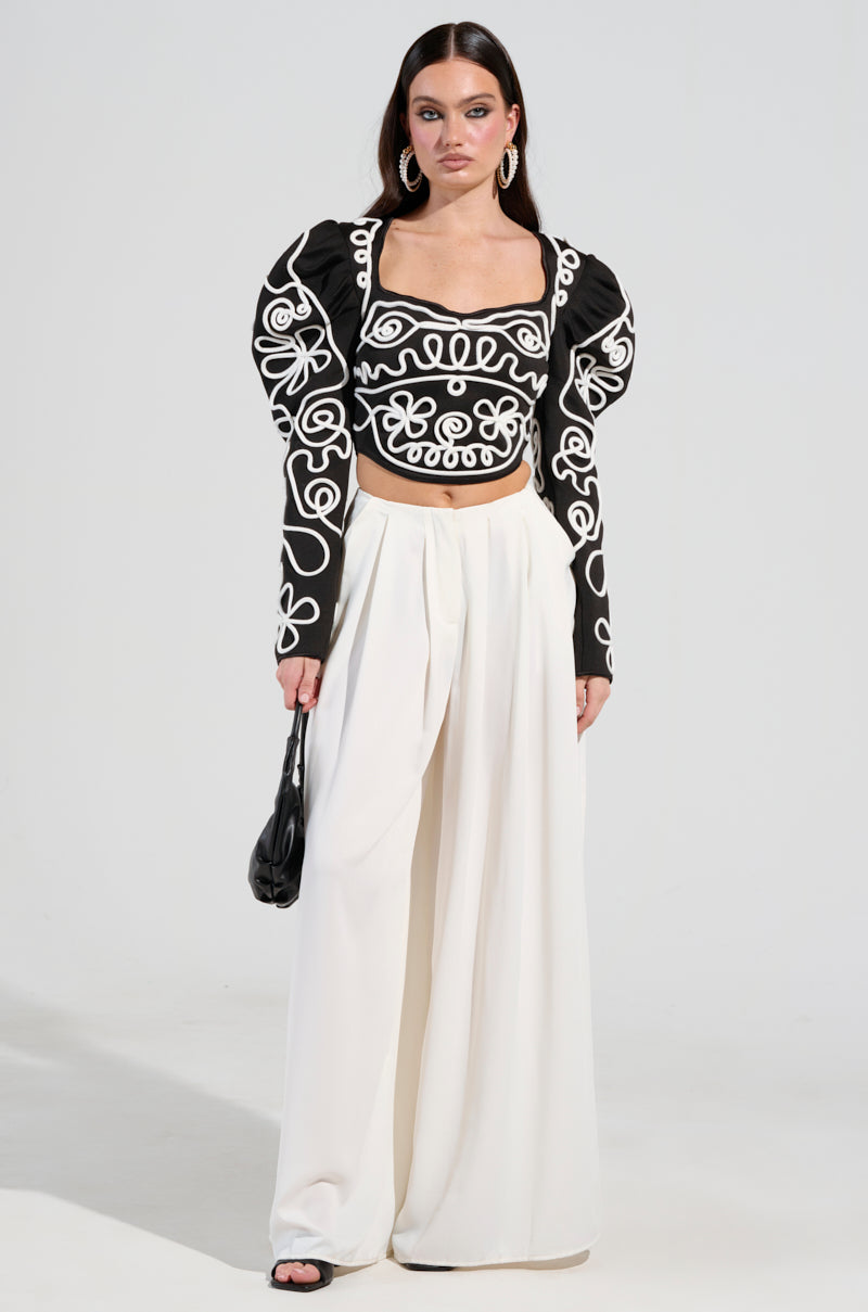 A woman stands wearing a BLURRED LINES LONG SLEEVE BLOUSE featuring abstract swirling patterns, complemented by wide-leg white trousers. She accessorizes with a small black handbag, her long straight hair flowing, and hoop earrings perfectly enhancing her stylish ensemble.